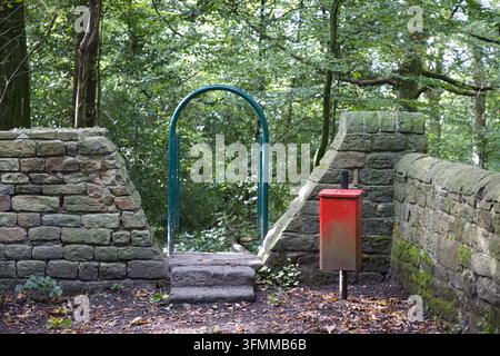Spreco di muro in pietra rustico con cancello ad arco verde acqua, gradini in pietra e cestino rosso nel bosco. Foto Stock