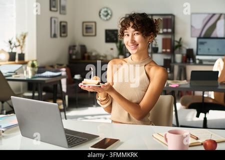 Ritratto di una donna biracial che tiene una torta di compleanno in un ufficio creativo Foto Stock