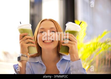 Donna sorridente solleva due bevande miscelate verdi ricoperte di panna montata mentre è in piedi davanti a finestre luminose e una vegetazione tropicale illuminata dal sole all'interno Foto Stock