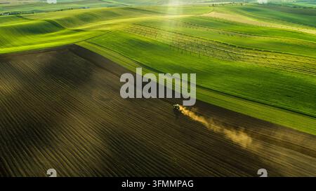 Vista aerea di un trattore che ara un campo nella paesaggio ondulato della Moravia, creando scie di polvere sotto la luce dell'ora dorata nel distretto di Brno-Città Foto Stock