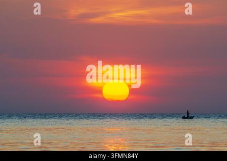Pescatore solitario in una piccola barca a remi davanti a un tramonto luminoso, il mare calmo e vasto, la silhouette dell'isola di Koh Chang nella luce calda Foto Stock