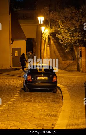 Auto parcheggiata e pedoni su una strada acciottolata illuminata da calde lampade tradizionali in un quartiere storico di notte. Foto Stock