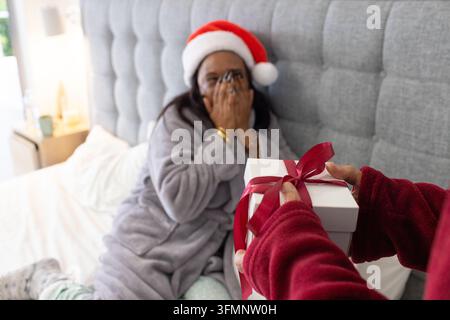 Uomo anziano diverso che offre una scatola regalo a una donna seduta sul letto che indossa una vestaglia grigia e un cappello di Natale Foto Stock