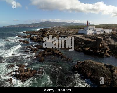 Vista aerea di un faro abbandonato accanto alla costa atlantica frastagliata del Portogallo, dove le onde si infrangono su scogli vulcanici scuri sotto un cielo parzialmente nuvoloso Foto Stock