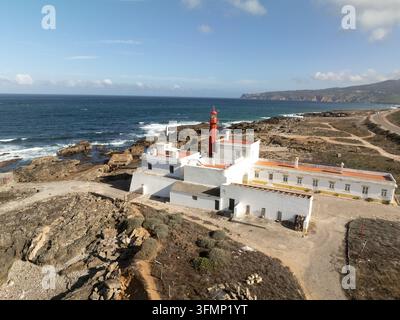 Vista aerea di un complesso di faro abbandonato sulla frastagliata costa atlantica del Portogallo, con edifici bianchi, una torre rossa, una costa rocciosa, onde Foto Stock