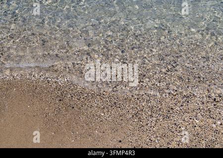 Acqua di mare chiara e poco profonda che si riversa delicatamente su una spiaggia di ciottoli e sabbia con piccole pietre visibili sotto la superficie trasparente creando una natura dettagliata Foto Stock