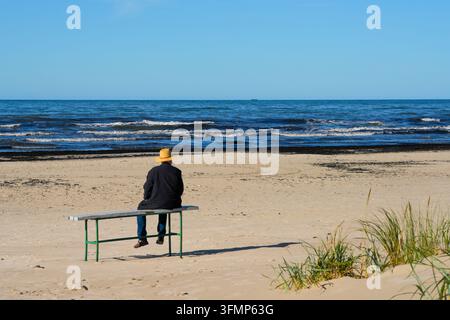 Uomo anziano seduto da solo su una panchina e che guarda il mare da una tranquilla spiaggia sabbiosa in autunno Foto Stock