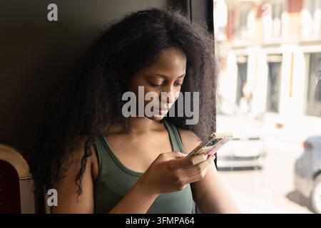 Una giovane donna con i capelli naturalmente ricci usa uno smartphone seduta vicino a una finestra, concentrata sullo schermo, con una luminosa scena urbana sullo sfondo Foto Stock