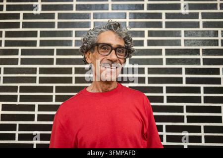 Ritratto di un uomo d'affari anziano sorridente con capelli grigi ricci e occhiali, che indossa una camicia rossa in piedi davanti a un muro di mattoni bianchi e neri Foto Stock