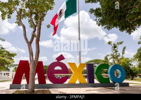 Insegna colorata del Messico sotto la bandiera nazionale messicana in un parco pubblico soleggiato, incorniciata da alberi a foglia, nuvole luminose e una piazza lastricata in Messico. Foto Stock