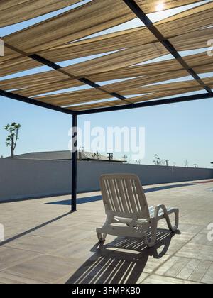 Una sedia a sdraio bianca si trova su una terrazza terrazzata sotto una struttura di pergola in tessuto, proiettando ombre a strisce nella luce del sole contro Foto Stock