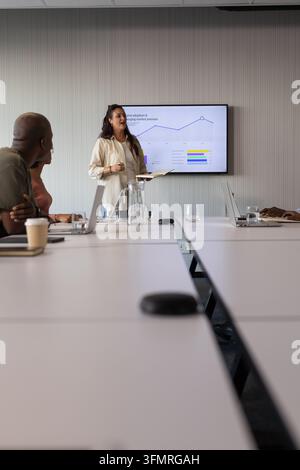 Squadra diversificata che ascolta mentre una donna indiana presenta in una sala conferenze con blocco note e schermo. Sala riunioni, presentazione, collaborazione, leadership Foto Stock