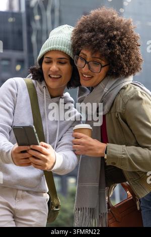 Diverse amiche si chinano, guardano uno smartphone in strada in città, tengono una tazza di caffè, un berretto. Urbano, inverno, felpa con cappuccio, giacca, occhiali, maglieria, sciarpa Foto Stock