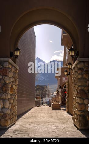 Canmore, Alberta, Canada - 02 12 2025: Vista attraverso un arco ricurvo in pietra e stucco lungo un vicolo stretto con pavimentazione decorata nel centro di Canm Foto Stock