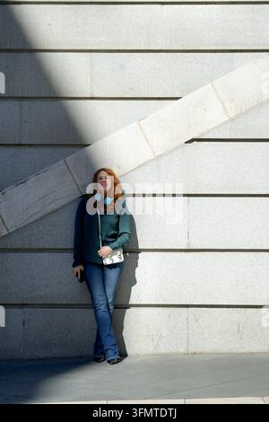 Donna elegante con cuffie in piedi all'aperto davanti a un muro di cemento geometrico, illuminata da una luce solare drammatica e ombre in una città contemporanea Foto Stock