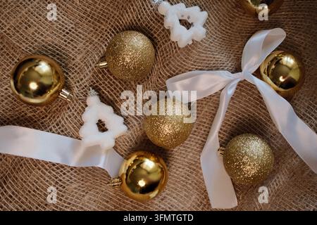 Flat lay di palline dorate di Natale, ornamenti a forma di fiocco di neve bianchi e un nastro delicato disposti su un tessuto strutturato, offrendo uno sfondo stagionale raffinato Foto Stock