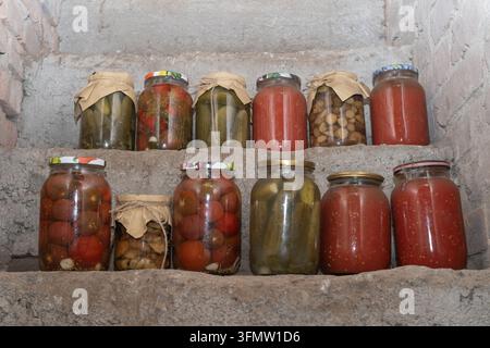 Assortiti verdure sottaceto fatte in casa, cetrioli e succo di pomodoro conservati in barattoli di vetro su scaffali all'interno di una cantina rustica. Foto Stock