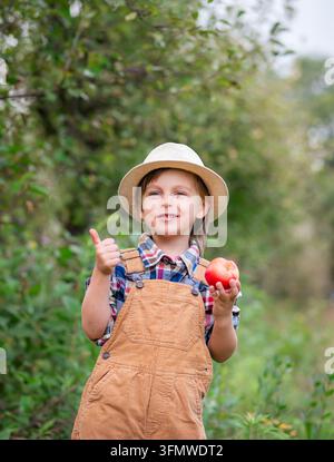 Ragazzo con il cappello che tiene in mano la mela rossa morsa e mostra i pollici nel frutteto durante la raccolta autunnale. Foto Stock