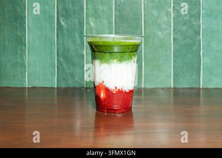 Bevanda stratificata alla fragola e matcha in una tazza di plastica contro un muro di legno verde Foto Stock