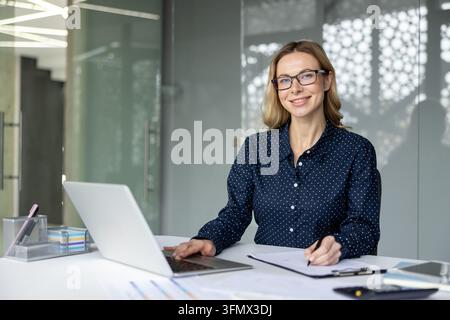 Donna d'affari sorridente con occhiali seduta alla scrivania, lavora attivamente al laptop e prende appunti importanti, dimostrando produttività e successo professionale Foto Stock