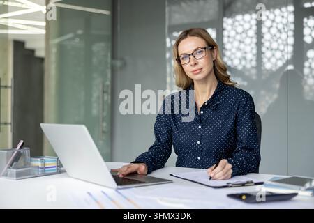 Donna d'affari professionale con occhiali e camicetta a pois a una scrivania moderna, scrive su un laptop e prende appunti tra documenti e grafici, concentrata sulla pianificazione e l'analisi Foto Stock