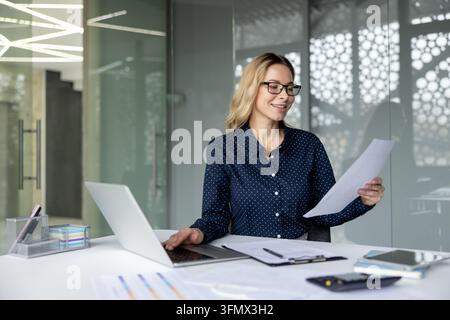 Donna d'affari con occhiali che esamina documenti e digita su un laptop in un ufficio moderno, concentrata e produttiva mentre gestisce compiti finanziari e di gestione con sicurezza Foto Stock