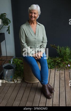 Donna anziana seduta su uno sgabello intrecciato in terrazza, indossa una camicia verde salvia e jeans, vicino a un annaffiatoio Foto Stock