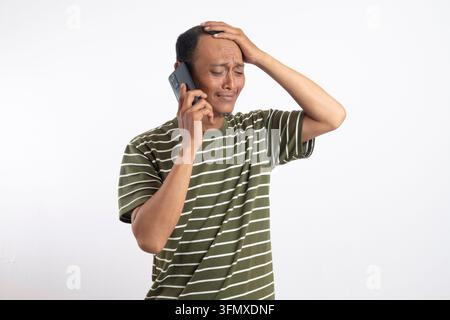 Un giovane uomo asiatico scioccato e confuso che si tiene la testa mentre usa uno smartphone per fare una chiamata, isolato su uno sfondo rosa. Foto Stock