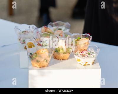 Vari antipasti serviti in piccole coppette di plastica trasparente su una superficie bianca durante un evento all'aperto. Foto Stock