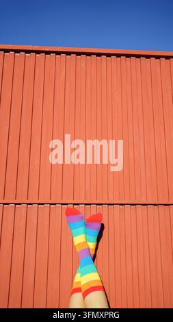 Gambe con calzini vivaci a righe arcobaleno appoggiati contro un muro ondulato arancione con cielo blu Foto Stock