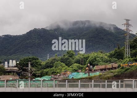Chongqing, Cina – 8 giu 2026 – La nebbia avvolge le rigogliose colline verdi in questo sereno paesaggio montano, con linee elettriche ad alta tensione che si ergono in alto Foto Stock