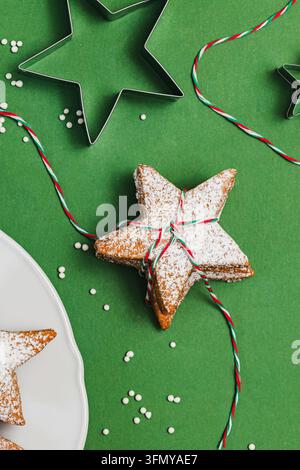 Pila di biscotti allo zenzero a forma di stella spolverati di zucchero e legati con uno spago rosso e bianco, con stampini per biscotti e zuccherini su verde. Foto Stock