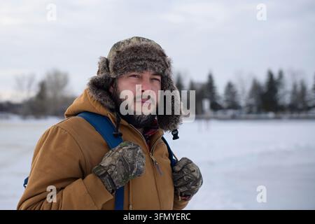 Scatto medio di un uomo barbuto pensieroso che indossa un cappello di pelliccia e una giacca invernale mentre fa escursioni accanto al lago Sylvan coperto di neve in Alberta, Canada, sotto un o Foto Stock