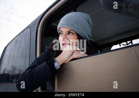Giovane donna pensierosa che indossa un berretto di lana grigio e una sciarpa appoggia il mento sulle mani giunte mentre guarda lontano dal finestrino di un veicolo fuoristrada nero Foto Stock
