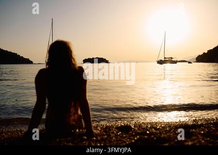 Silhouette di una donna seduta su una spiaggia che osserva barche a vela all'ora dorata del tramonto su un mare calmo Foto Stock