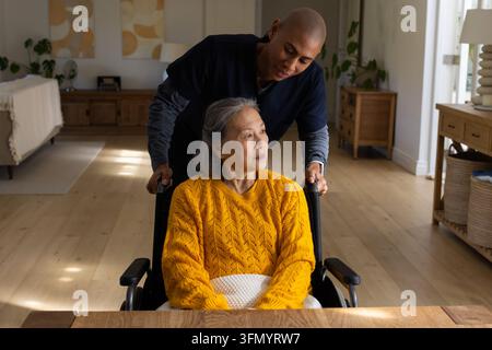 Donna anziana cinese seduta su una sedia a rotelle in salotto, coperta di maglia in grembo, badante che spinge Foto Stock