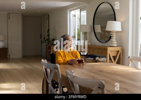 Donna anziana asiatica con maglione senape seduta su una sedia a rotelle al tavolo da pranzo mentre tiene una tazza da tè Foto Stock