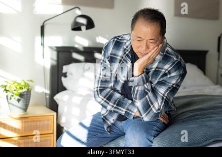 Uomo asiatico anziano seduto sul letto, indossa una camicia a quadri, appoggia la guancia sulla mano, la luce del sole che entra Foto Stock