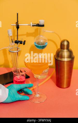 Una mano guantata tiene un bicchiere coupé di cocktail arancione accanto a vetreria da laboratorio, liquidi colorati, tubi e uno shaker in ottone su uno sfondo giallo vivace Foto Stock