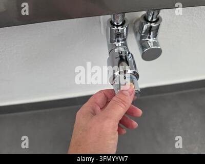 Mano che regola la valvola termostatica sui tubi cromati sotto il radiatore bianco, primo piano del controllo della temperatura del sistema di riscaldamento domestico all'interno Foto Stock