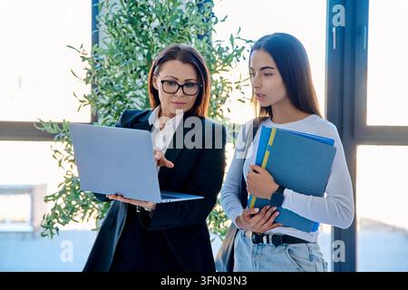 Insegnante che mostra lo schermo del laptop a uno studente che tiene cartelle vicino alla finestra in un ufficio moderno Foto Stock
