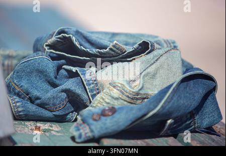 Una fotografia orizzontale in primo piano che cattura una classica giacca di jeans blu sbiadita lasciata in un mucchio accartocciato su una panchina di legno verde consumata sotto brig Foto Stock