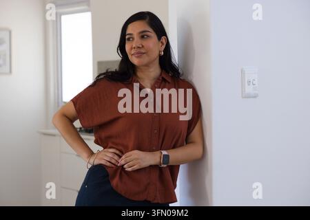 Donna di mezza età appoggiata a un muro bianco a casa, indossa una camicetta ruggine, smartwatch, braccialetto, orecchini Foto Stock