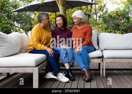 Amiche diverse, una anziana, che chiacchierano su un divano all'aperto sul ponte sotto un ombrellone, indossando maglie Foto Stock