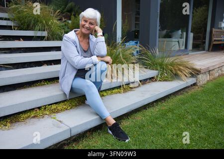 Donna anziana seduta sui gradini di cemento di una casa moderna, indossa abiti sportivi e controlla l'orologio da polso Foto Stock