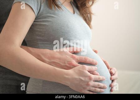 Donna incinta che culla il suo ventre, abbracciata da dietro dal partner. Madre in attesa e marito che condividono un momento tenero in studio con sfondo neutro. Futura mamma nel suo primo viaggio di gravidanza Foto Stock