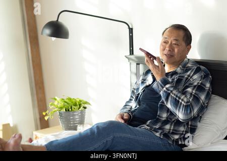 Uomo asiatico anziano seduto sul letto in camera da letto, tiene uno smartphone vicino alla bocca, indossa una flanella Foto Stock