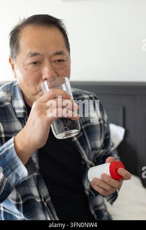 Uomo asiatico anziano in camicia a quadri seduto sul letto, che tiene una bottiglia con tappo rosso e beve da un bicchiere trasparente Foto Stock