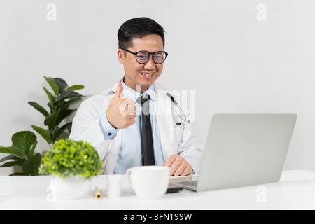 Giovane medico maschio sorride, dà il pollice in su mentre lavora su un laptop. Indossa occhiali, camice bianca e stetoscopio. Una pianta verde e una tazza sono accanto al laptop Foto Stock