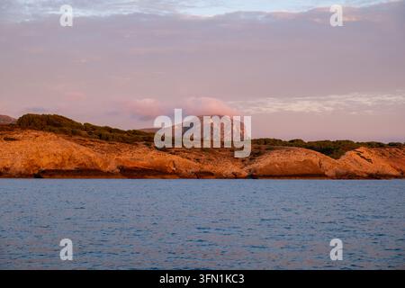 La luce calda dell'ora dorata avvolge scogliere rosse frastagliate e un picco montuoso drammatico sulla costa di Maiorca, ideale per concetti di viaggio e natura. Foto Stock